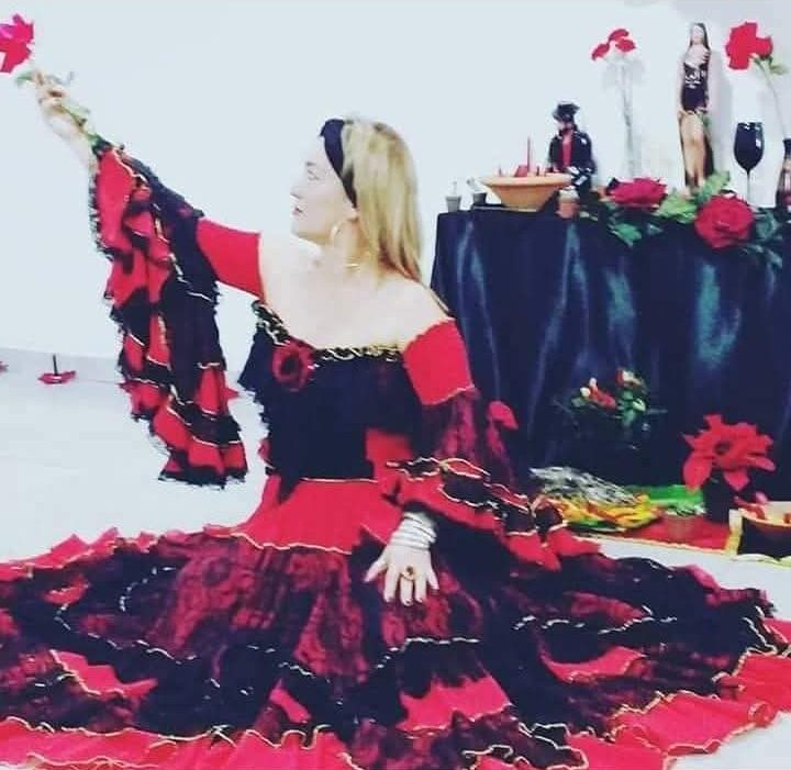 Kelli Garcia com vestido vermelho e preto de babados, erguendo uma rosa diante de um altar